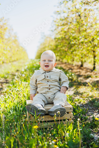 A cute baby boy is sitting on the grass in the park and smiling. A stylish baby in a beige romper.