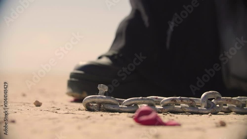 A woman in black is hammering a nail to fix a chain on dry cracked soil of a desert. A metaphor of restriction, a symbol of restraint and burden.
