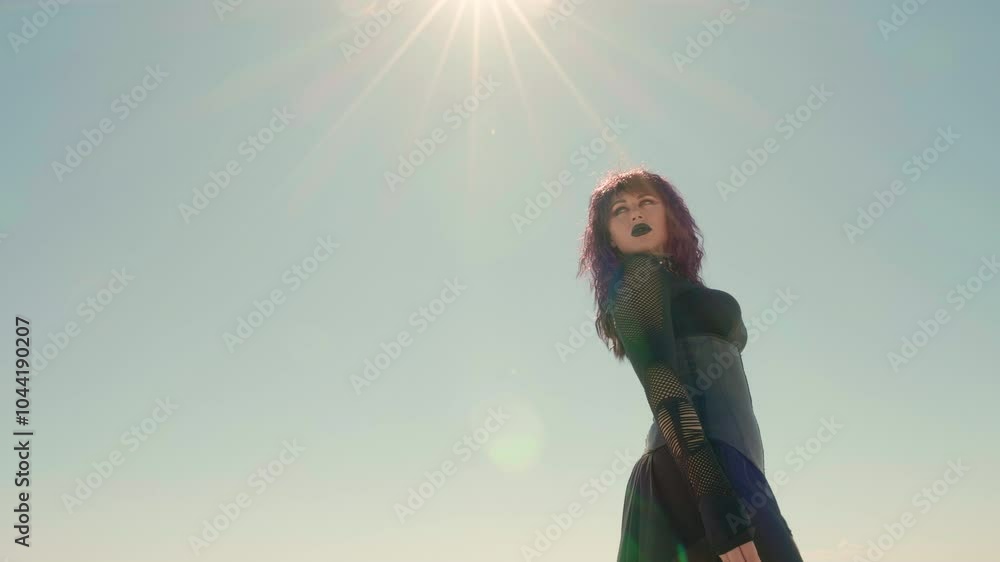 Low angle shot of a young Goth woman, with pale sky and sun flare in the background. Gothic girl strides through the barren landscape with her head turned back, as if looking at something left behind. Low angle shot of a young Goth woman, with pale sky and sun flare in the background. Gothic girl strides through the barren landscape with her head turned back, as if looking at something left behind.