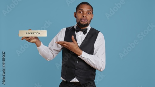 Hotel concierge staff showing the reception indicator at front desk, greeting guests at five star holiday resort in studio. Skilled professional bellboy holds sign against blue background. Camera B. photo