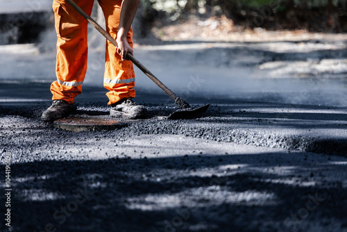 Worker repairing asphalt road surface on a sunny day using a smooth lute rakein an urban area with trees nearby