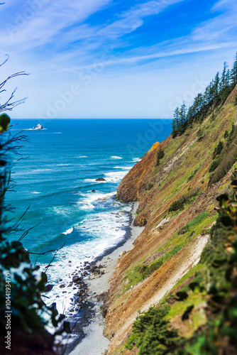 Rugged coastline stretches along the ocean, with steep cliffs covered in lush greenery. Waves gently crash against the rocky shore under a vibrant blue sky. Viewed from Clatsop Trail, Oregon.