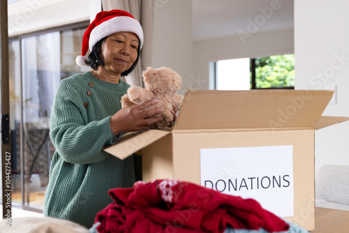 Senior asian senior woman in Santa hat packing teddy bear into donation box at home, Christmas time photo