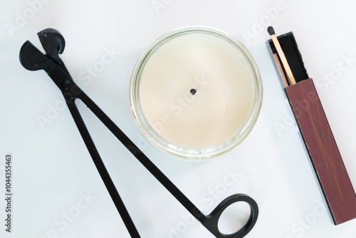 Scented candle in a glass jar with a candle trimmer and a matchstick over a White background.