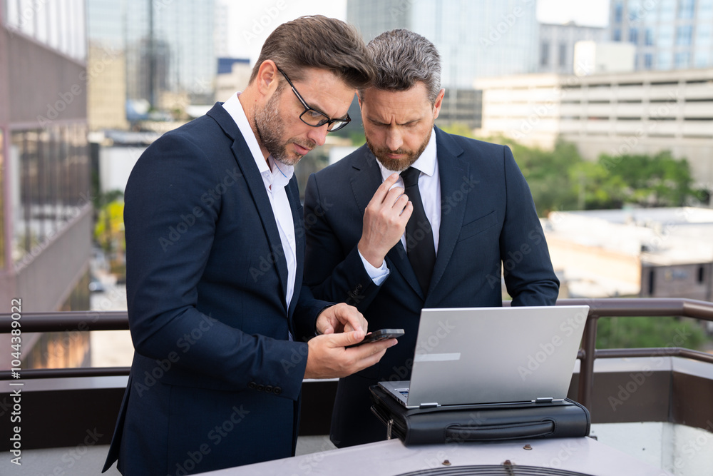 Two businessman on the street, deeply engrossed, eyes fixed on their laptop screen. Two handsome young businessmen in classic suits using laptop. Business men team using laptop outdoor. Business man Two businessman on the street, deeply engrossed, eyes fixed on their laptop screen. Two handsome young businessmen in classic suits using laptop. Business men team using laptop outdoor. Business man