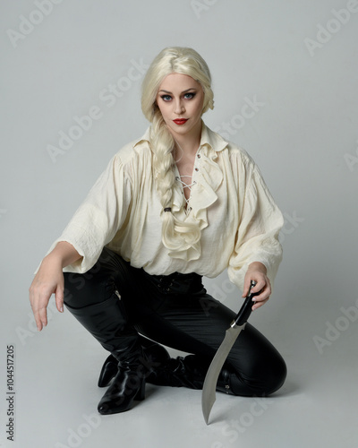 full length portrait of beautiful female model, wearing vintage gothic fantasy ruffle blouse white shirt, leather pants, blonde hair. holding knife weapon. sitting pose, isolated on studio background. photo