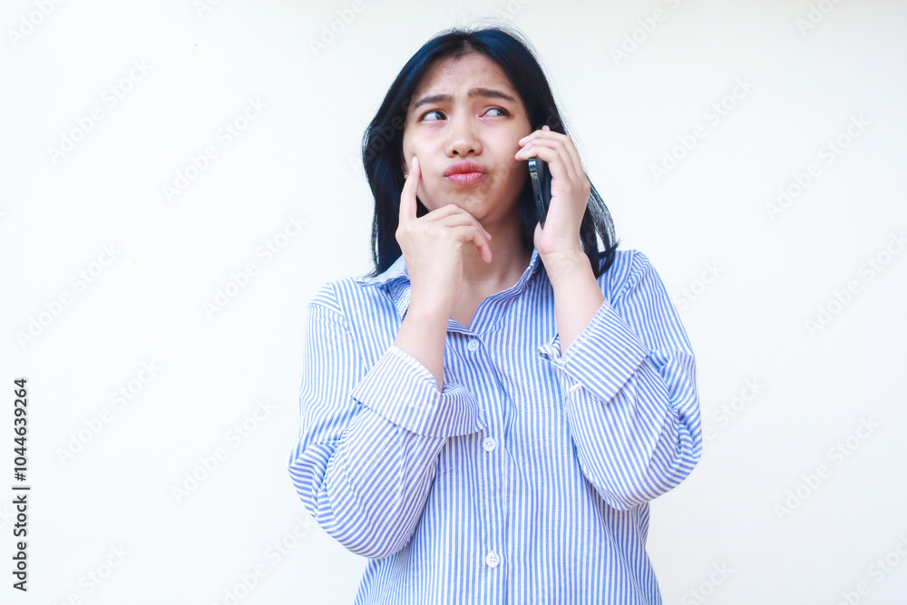thoughtful asian woman using smartphone with hands on cheek thinking problem wearing blue striped casual shirt standing over isolated white background, look away thoughtful asian woman using smartphone with hands on cheek thinking problem wearing blue striped casual shirt standing over isolated white background, look away