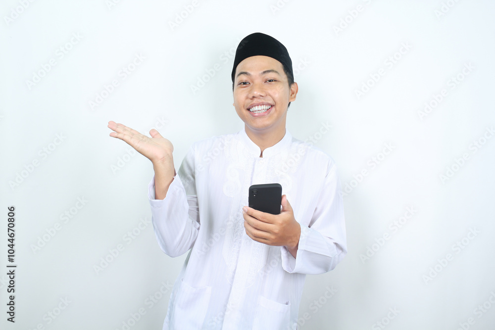pleased asian muslim man smiling while presenting hand to beside with holding mobile phone isolated pleased asian muslim man smiling while presenting hand to beside with holding mobile phone isolated