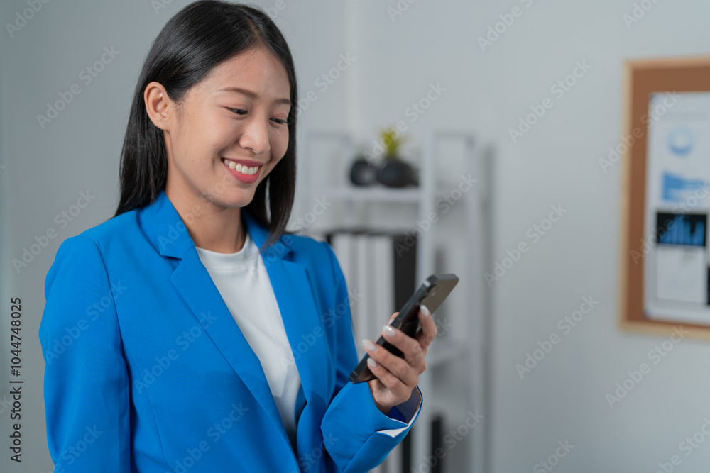 Young asian businesswoman is standing in her office, smiling while using her smartphone. She is wearing a blue blazer and has a professional demeanor Young asian businesswoman is standing in her office, smiling while using her smartphone. She is wearing a blue blazer and has a professional demeanor