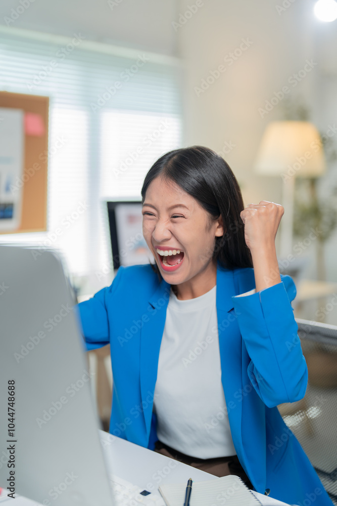 Young businesswoman is sitting at her desk raising her fist in the air, excited about what she is seeing on her computer screen Young businesswoman is sitting at her desk raising her fist in the air, excited about what she is seeing on her computer screen