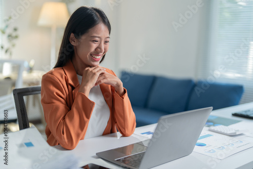 Asian businesswoman happily works from her home office, smiling at her laptop screen while tackling tasks related to business, finance, and sales
