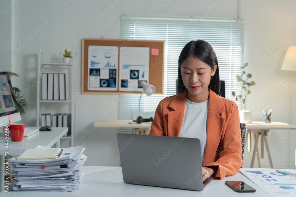 Young asian businesswoman is working from her home office. She is using a laptop computer and has a pile of paperwork on her desk Young asian businesswoman is working from her home office. She is using a laptop computer and has a pile of paperwork on her desk