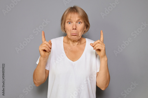 Frustrated disappointed senior woman wearing white T-shirt pointing up with displeased expression at empty advertising area against gray background raising her arms in disappointment