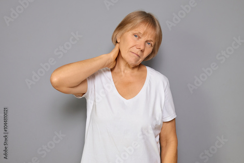 Worried mature woman wearing white T-shirt feeling pain in her neck and back showing signs of physical stress and anxiety dealing with her acute injury standing against gray background