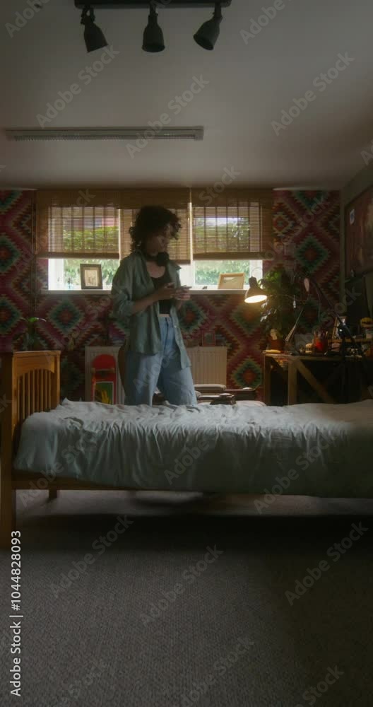 A young woman in casual clothes uses a mobile phone at home with a serious look. She sits down on the bed A young woman in casual clothes uses a mobile phone at home with a serious look. She sits down on the bed