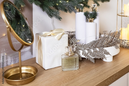 arranged holiday display wrapped gift in silver paper decorative glass perfume bottle and lush greenery. round white tray holds shimmering decorative items alongside. golden mirror. dressing tables 