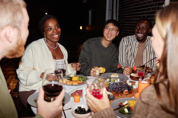 Diverse group of smiling adult people enjoying dinner party outdoors and celebrating holidays together, shot with flash