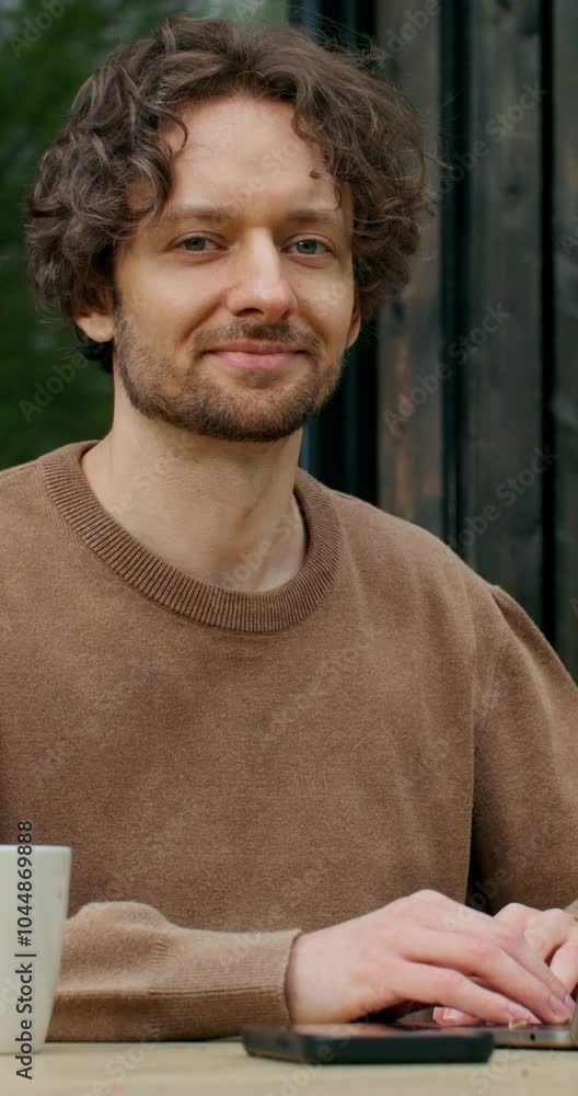 A man in casual clothes smiles looking at the camera sitting in front of an open laptop on the terrace of a stylish cottage on a summer day A man in casual clothes smiles looking at the camera sitting in front of an open laptop on the terrace of a stylish cottage on a summer day