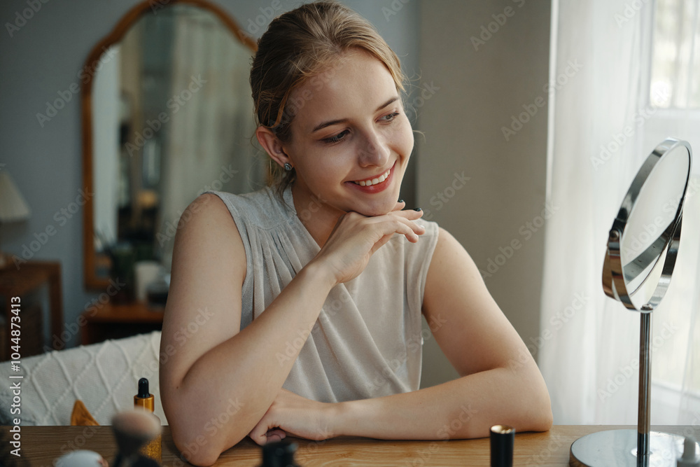 Young woman with light hair smiling and resting chin on hand, gazing at her reflection in small round mirror while sitting at vanity with makeup items scattered Young woman with light hair smiling and resting chin on hand, gazing at her reflection in small round mirror while sitting at vanity with makeup items scattered