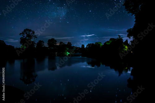 Starry night sky over the river and its reflection in the still water. The reflection of the silhouette of farmhouse and trees in the river at night.