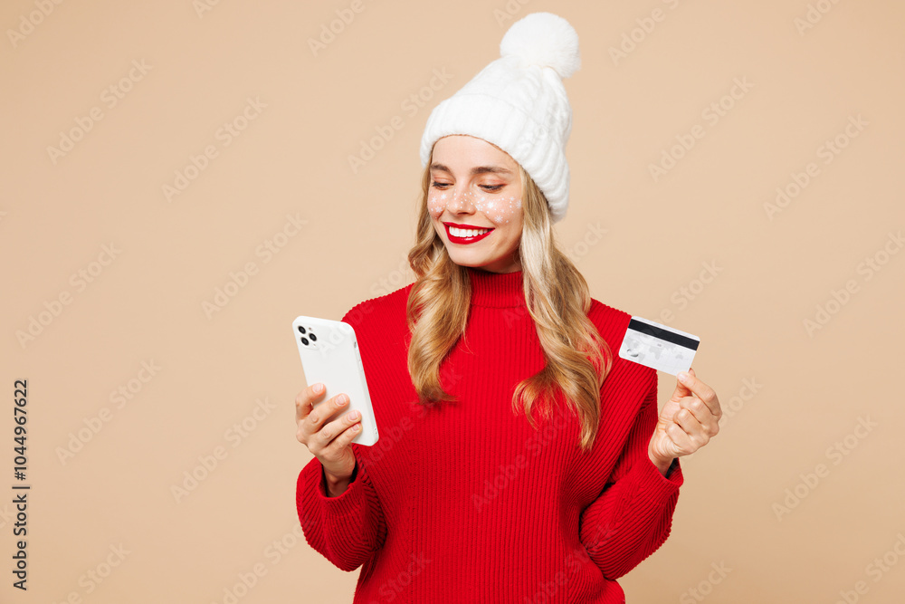 Merry young woman wearing red sweater hat posing using mobile cell phone hold credit bank card shopping online isolated on plain beige background. Happy New Year celebration Christmas holiday concept. Merry young woman wearing red sweater hat posing using mobile cell phone hold credit bank card shopping online isolated on plain beige background. Happy New Year celebration Christmas holiday concept.