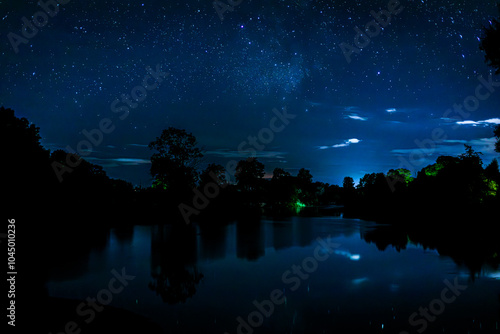 Starry night sky over the river and its reflection in the still water. The reflection of the silhouette of farmhouse and trees in the river at night.