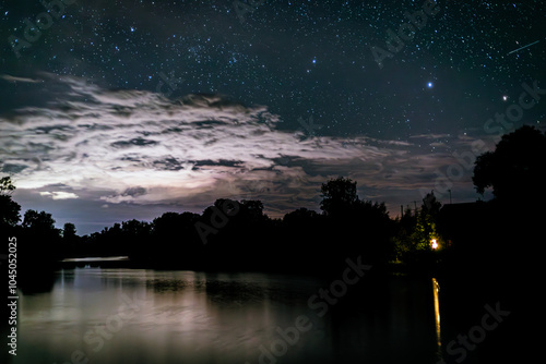 Starry night sky over the river and its reflection in the still water. Night clouds and a shooting star are visible in the sky