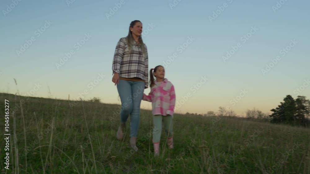 A mother and daughter walking hand in hand through a grassy field at sunset, with trees in the background. A mother and daughter walking hand in hand through a grassy field at sunset, with trees in the background.