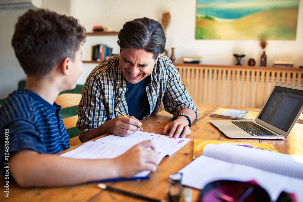 Father laughing while helping son with homework on desk Father laughing while helping son with homework on desk
