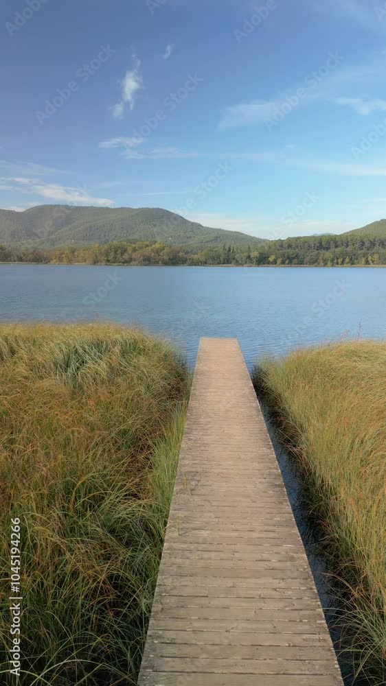 a wooden boardwalk extending through tall grass towards a calm lake. The lake is surrounded by forested hills and mountains in the background. a wooden boardwalk extending through tall grass towards a calm lake. The lake is surrounded by forested hills and mountains in the background.