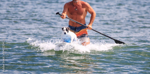 Man kneeling on a sufboard behind his dog catching a wave photo