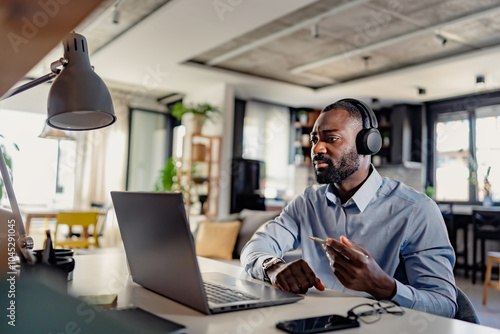 African-American businessman wearing headphones, engaged in a video call on his laptop in a modern home office. Focused and expressive during a virtual meeting.