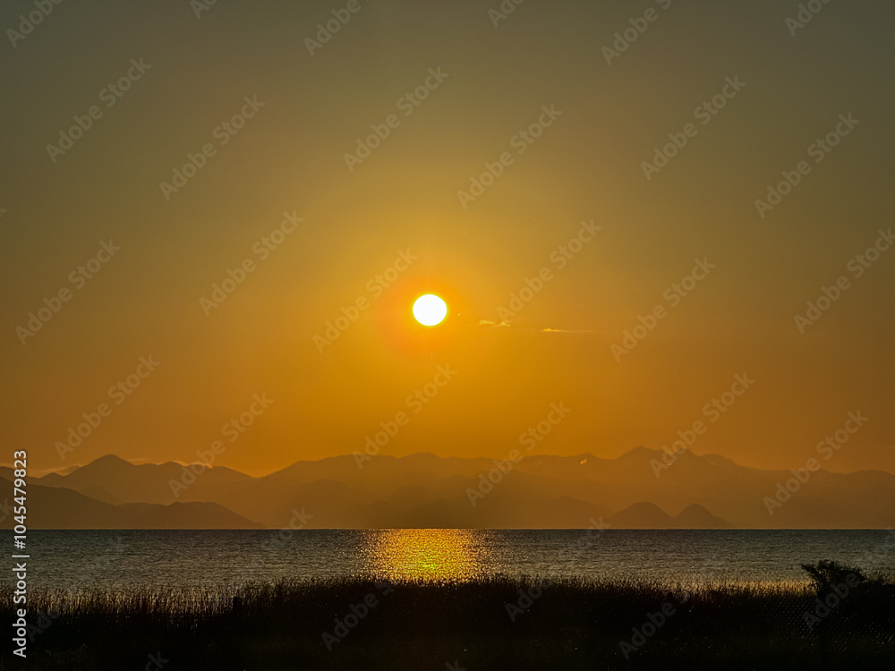 Breathtaking sunset over calm Skadar lake, Albania. Sun is setting behind Dinaric Alps mountains in Montenegro. Water reflects vibrant colors of sky creating shimmering path across the surface Breathtaking sunset over calm Skadar lake, Albania. Sun is setting behind Dinaric Alps mountains in Montenegro. Water reflects vibrant colors of sky creating shimmering path across the surface