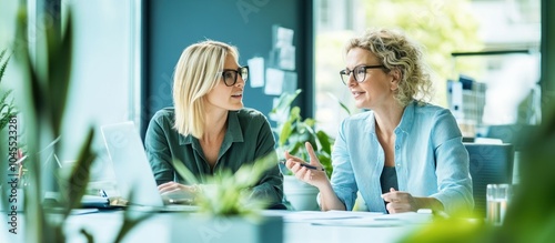 Two businesswomen discussing work at their desk in a modern office with green plants in the foreground.