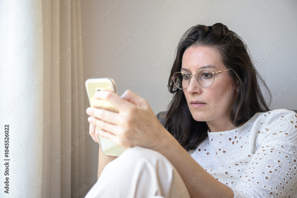 A woman with glasses looks intently at her smartphone, concentrated and absorbed. Sitting indoors near a curtain, she appears immersed in the screen, appearing calm and focused. A woman with glasses looks intently at her smartphone, concentrated and absorbed. Sitting indoors near a curtain, she appears immersed in the screen, appearing calm and focused.