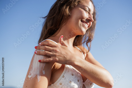 A happy and smiling young woman applies lotion, sunscreen on her shoulder as a hydration, health and skin care routine. Girl smiles, dermatology or female person with spf cream. Close up, copy space.