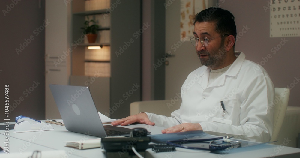 The doctor is talking on a video chat on a laptop sitting at a table in the medical office at the clinic The doctor is talking on a video chat on a laptop sitting at a table in the medical office at the clinic