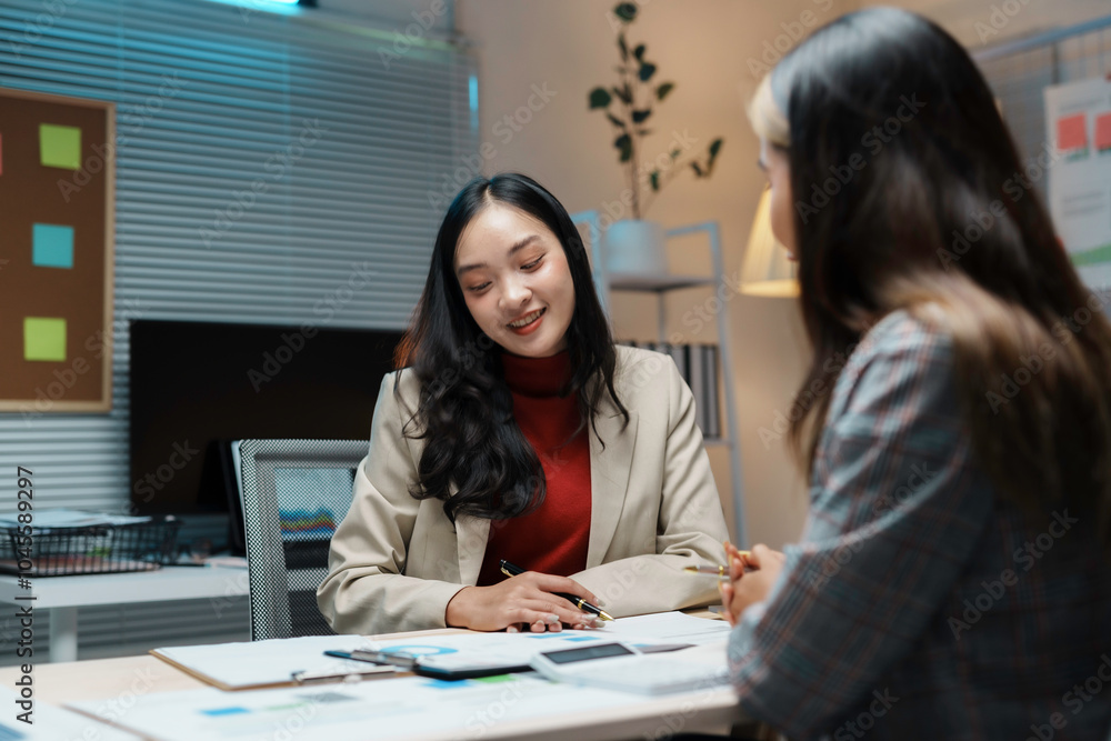 Two businesswomen are working late in a modern office, discussing financial documents and analyzing charts Two businesswomen are working late in a modern office, discussing financial documents and analyzing charts