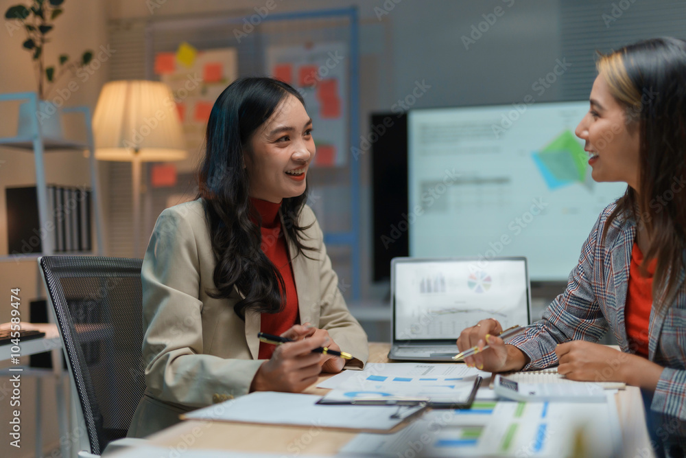 Two businesswomen smiling and talking about work while sitting at a desk in the office at night, analyzing financial data and planning a new project Two businesswomen smiling and talking about work while sitting at a desk in the office at night, analyzing financial data and planning a new project