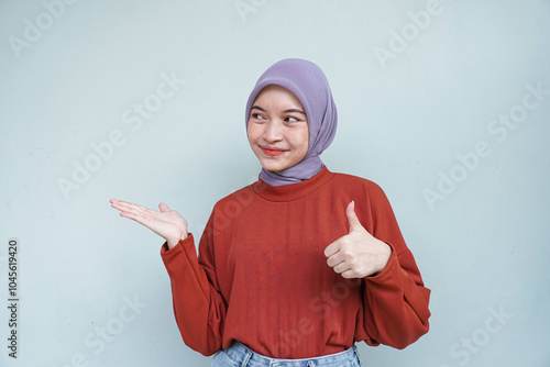 Happy young Asian Muslim woman wearing hijab presenting copy space beside her and thumbs up gesture. Isolated white background.