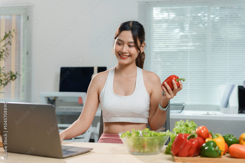 Young woman is smiling while holding a red bell pepper and working on a laptop with a bowl of fresh vegetables in front of her Young woman is smiling while holding a red bell pepper and working on a laptop with a bowl of fresh vegetables in front of her