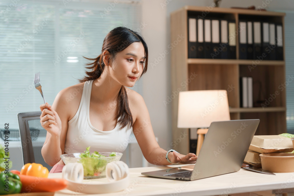 Young asian woman is enjoying a healthy salad while working on her laptop in her home office Young asian woman is enjoying a healthy salad while working on her laptop in her home office