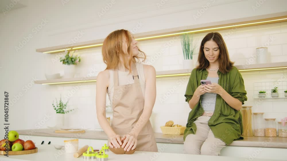 Lesbian couple in love is preparing for Christmas and preparing gingerbread cookies in the kitchen wearing aprons. Red-haired girl makes dough and brunette uses smartphone Lesbian couple in love is preparing for Christmas and preparing gingerbread cookies in the kitchen wearing aprons. Red-haired girl makes dough and brunette uses smartphone