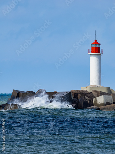 Small white lighthouse with red top and roof, wave washed concrete block breakwater of city port.