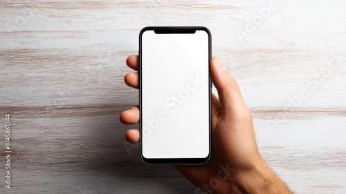 A person holding a blank smartphone against a light wooden background in a well-lit setting during the day with focus on the device's screen