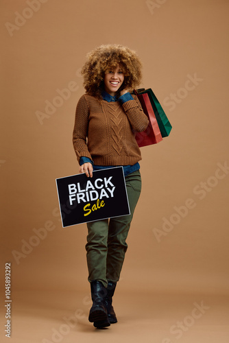 Happy mixed race woman holding shopping bags and Black Friday sale placard while looking at camera. photo