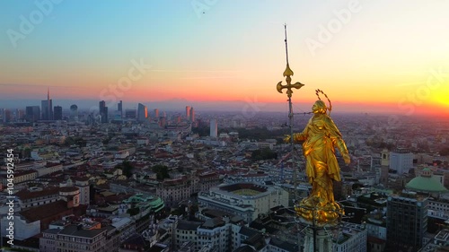 Aerial view of the statue of the Madonna on the central spire of the Duomo and the city's cathedral at dawn. Roofs of houses and skyscrapers. Sun over the horizon. duomo square. Italy Milan 11.04.2025