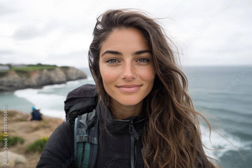 A woman with long hair stands on a cliffside overlooking the ocean, wearing a black coat and carrying a backpack, radiating calmness in the cloudy weather. A woman with long hair stands on a cliffside overlooking the ocean, wearing a black coat and carrying a backpack, radiating calmness in the cloudy weather.