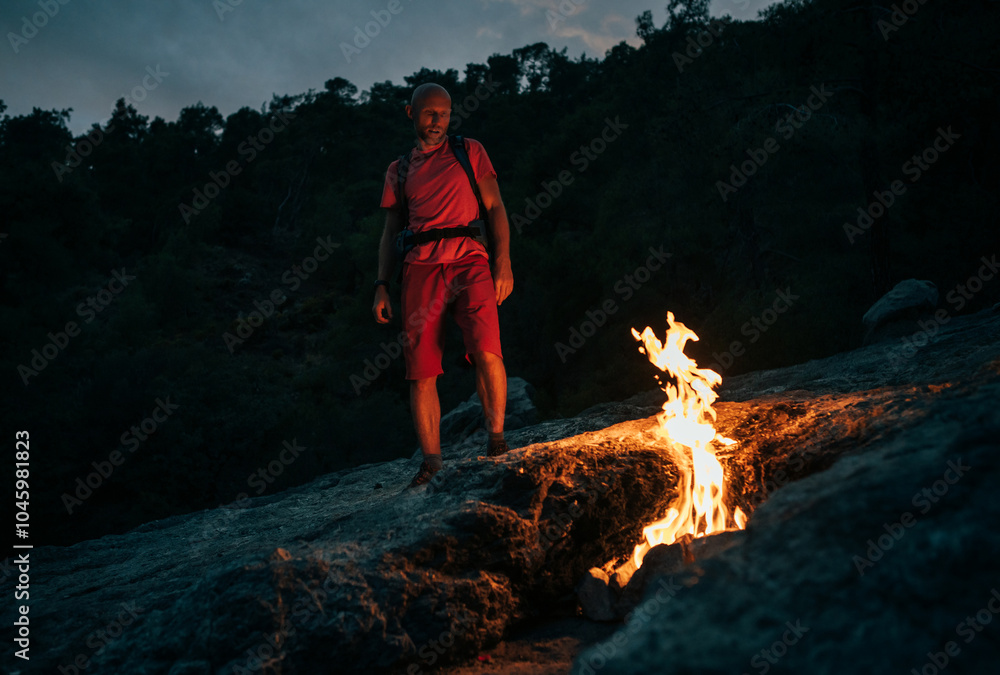 Obraz premium Hiker with backpack sits beside natural flames of Yanartaş, also known as eternal fire of Mount Chimaera, in Turkey. The night scene captures the unique fire phenomenon emerging from the mountain