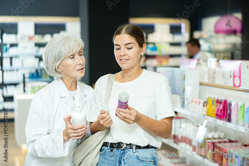 Senior woman pharmacist helps girl buyer to decide on choice of intimate lubricant gel. At pharmacy, female chemist shows lubricant and convinces doubter to purchase mini version of care product.